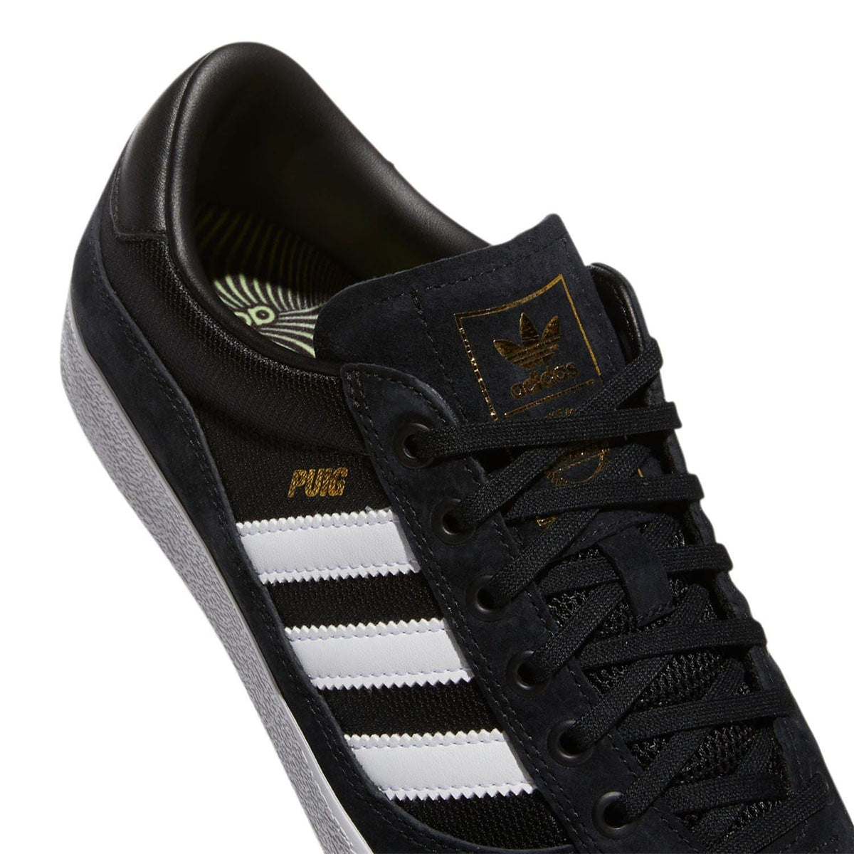 Black suede indoor trainer close-up with white stripes, laces, and gold badge details.