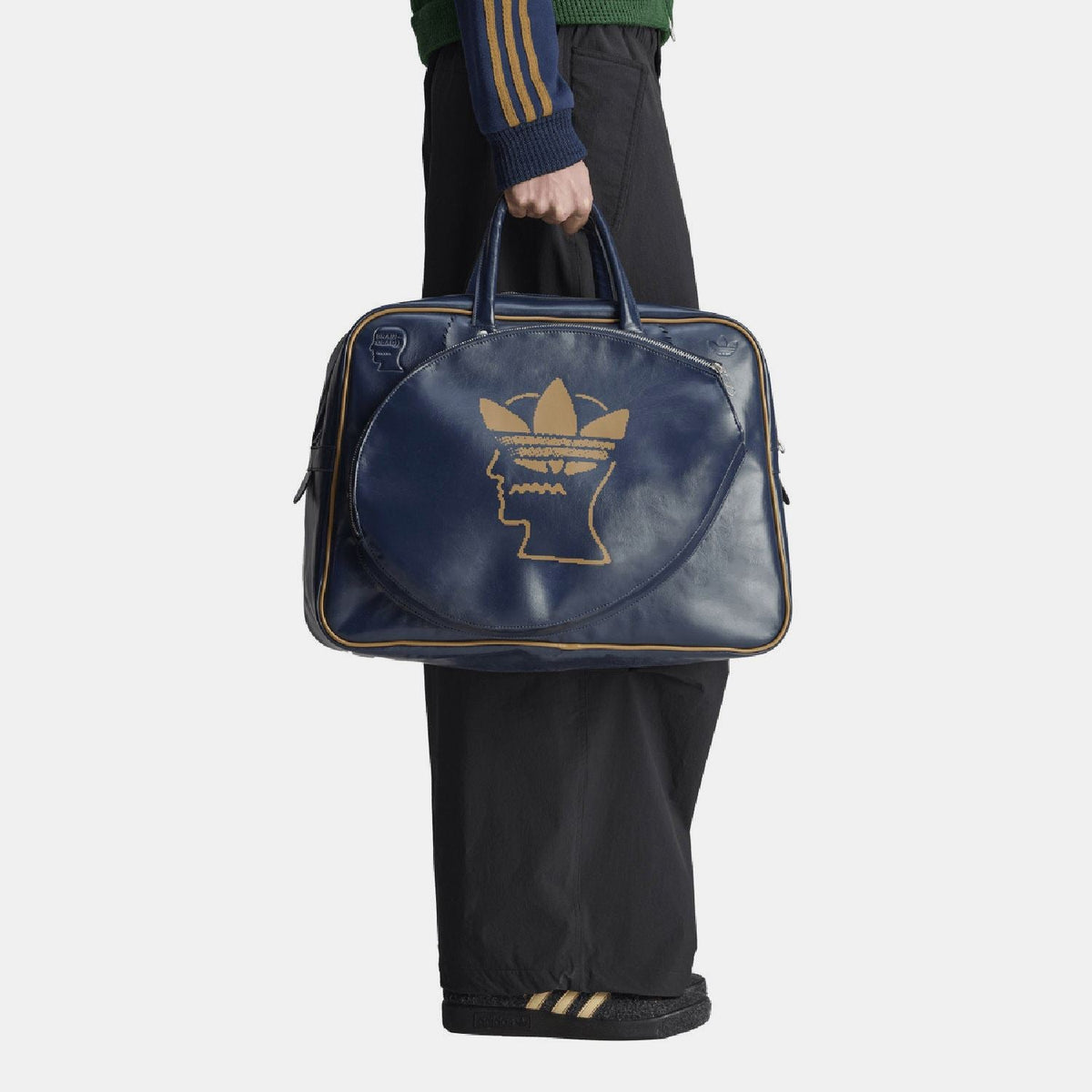 Navy duffel bag held in hand with front gold Trefoil head logo and side zipper ends.