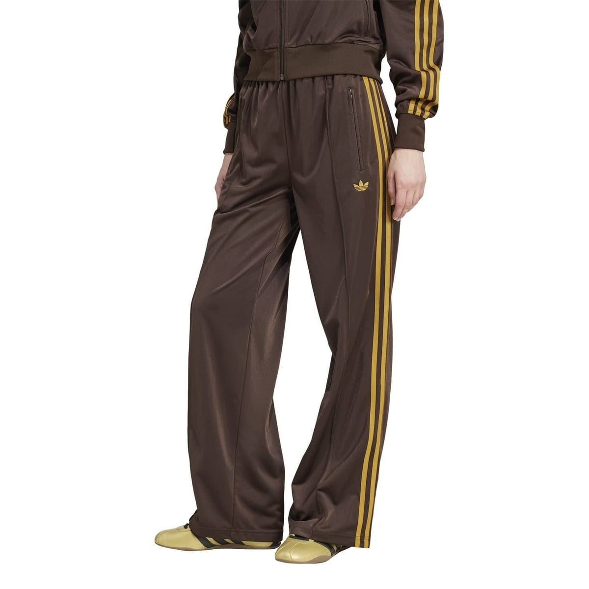 Brown adidas Firebird track pants with gold side stripes and trefoil logo, loose fit