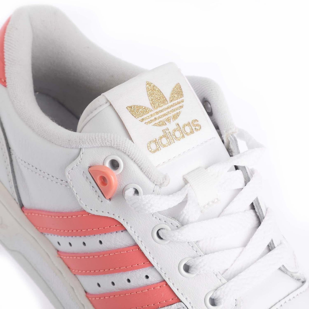 White adidas Rivalry Low sneaker close-up with gold adidas logo and coral lace tab